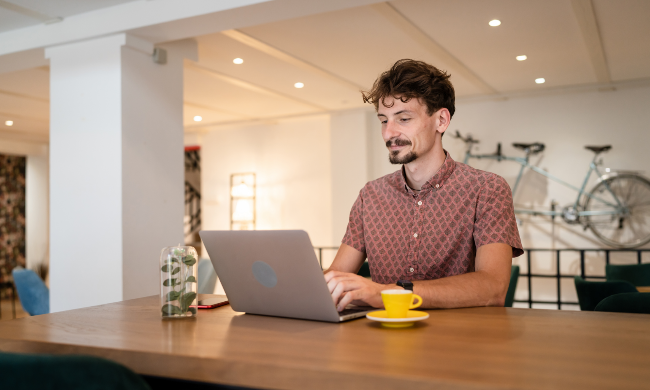 Man typing on a laptop. A yellow espresso cup and saucer sits on a table to his left.